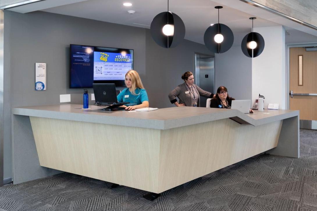 Two Kettering employees and a student worker  work at computers at the Library Services desk in the Learning Commons